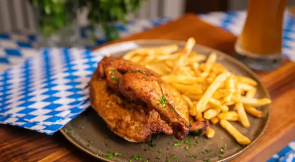 Ein knuspriger poulet roti mit Pommes Frites, garniert mit Kräutern, auf einem Teller mit blau-weiß karierter Serviette und einem Glas Bier.