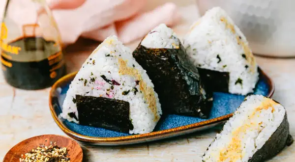 Drei dreieckige Onigiri mit Thunfisch-Mayo-Füllung, umhüllt von Nori-Algen, auf einem blauen Teller. Daneben eine Flasche Sojasauce und eine Schale mit Gewürzen.