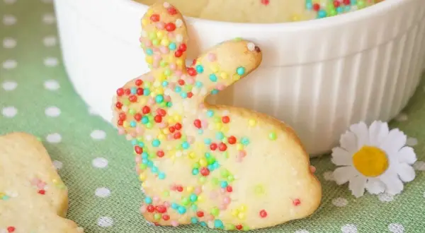 Bunte Osterkekse zum Ausstechen in Hasenform, verziert mit bunten Streuseln, liegen auf einem grünen Tupfentuch neben einer weißen Schüssel.