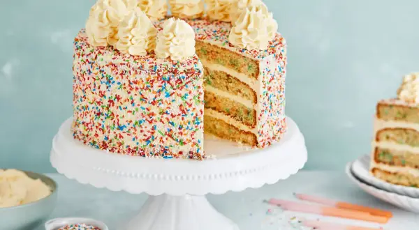Eine leckere Torte mit weißem Zuckerguss und bunten Streuseln, gekrönt mit Sahnetuffs. Ein Stück ist angeschnitten und zeigt die saftigen Schichten.