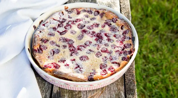 Ein frisch gebackener Clafoutis aux cerises, bestäubt mit Puderzucker, auf einem rustikalen Holztisch im Grünen.