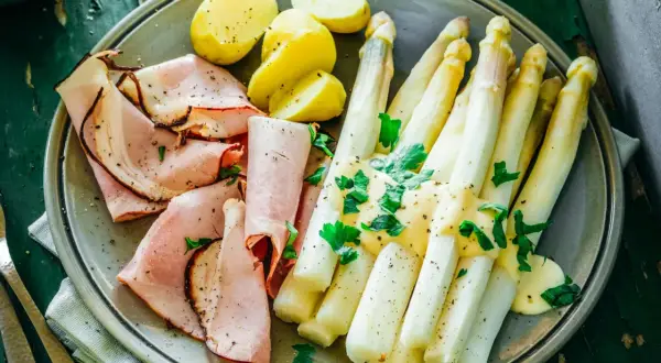 Ein Teller mit gekochtem Spargel, Schinken und Kartoffeln, garniert mit Petersilie. Perfekt für die besten Spargelrezepte!