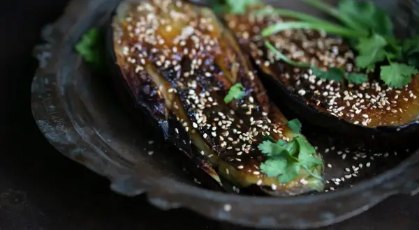 Zwei Hälften von gegrillter Miso Aubergine, bestreut mit Sesam und garniert mit Koriander, auf einem rustikalen Teller.
