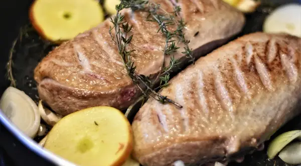Zwei gebratene Entenbrüste mit Rosmarin, Apfelscheiben und Zwiebeln in einer Pfanne. Ein köstliches entenfilet rezept.