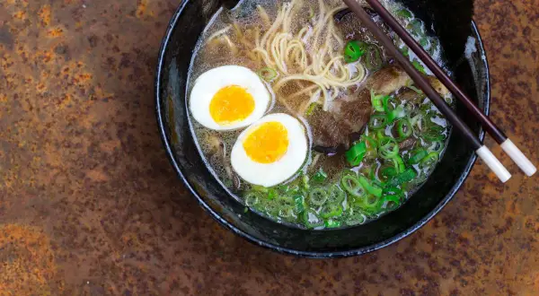 Schüssel mit dampfendem Ramen, zwei halbierten Eiern, Nudeln, Fleisch und Frühlingszwiebeln. Perfekt, um Ramen selbst zu machen!