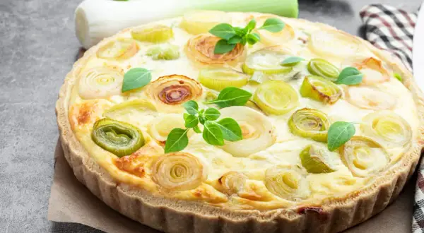 Herzhafte Quiche aux poireaux mit goldbraunem Teig und frischen Lauchringen, garniert mit grünen Kräutern.