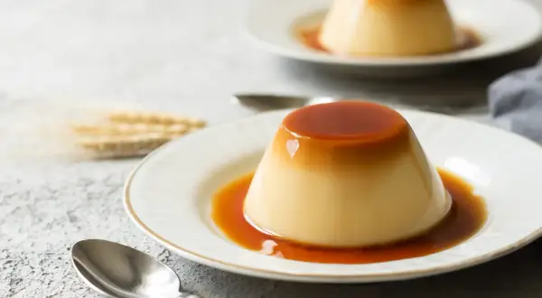 Zwei cremige flan kuchen mit flüssiger Karamellsoße auf weißen Tellern, daneben Löffel und ein paar Ähren.