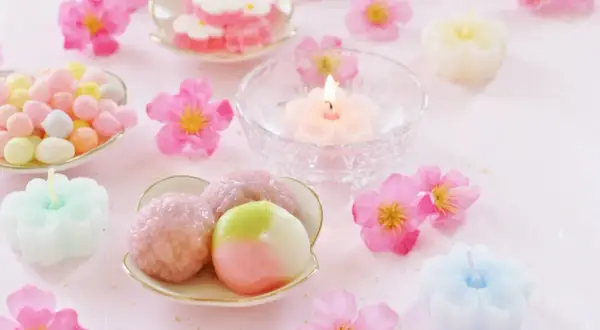 Zarte japanische Desserts, bunte Süßigkeiten und blütenförmige Kerzen auf rosa Untergrund.