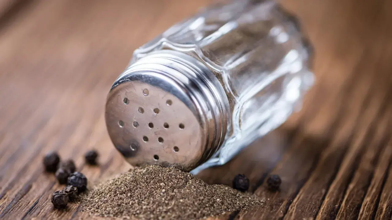 Ein Glasstreuer mit Metallaufsatz liegt auf einem Holztisch. Gemahlener Pfeffer und ganze Körner sind daneben verstreut. So kann man den Pfeffer neutralisieren.