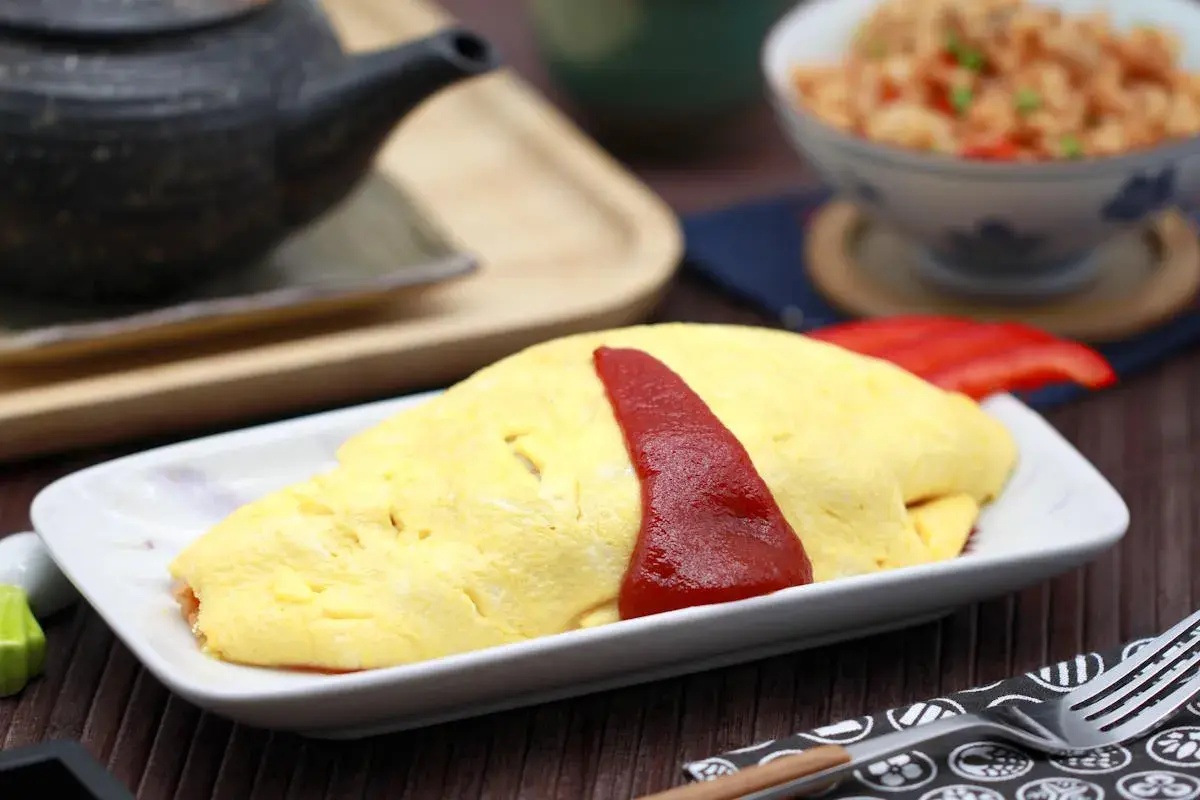 Ein leckeres Omurice Rezept: goldgelbes Omelett mit Ketchup-Streifen auf einem Teller, daneben eine Schüssel mit gebratenem Reis.