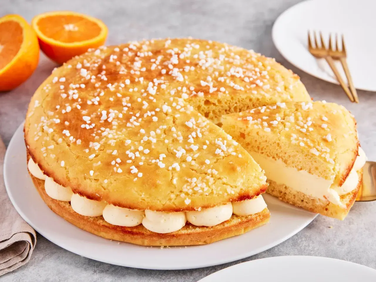 Eine köstliche Tarte Tropezienne, bestreut mit Zuckerperlen, gefüllt mit cremiger Vanillecreme. Daneben liegen Orangen.