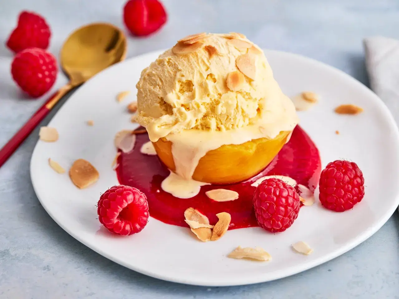 Ein Pfirsich Melba Rezept Original: Ein Pfirsich mit Vanilleeis, Himbeersauce und Mandelsplittern auf einem Teller, garniert mit frischen Himbeeren.