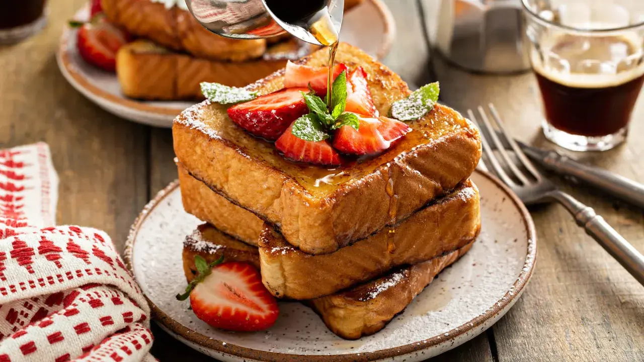 Ein Stapel goldbrauner Pain Perdu, garniert mit Erdbeeren und Minze, wird mit Sirup übergossen. Daneben ein Glas Kaffee.