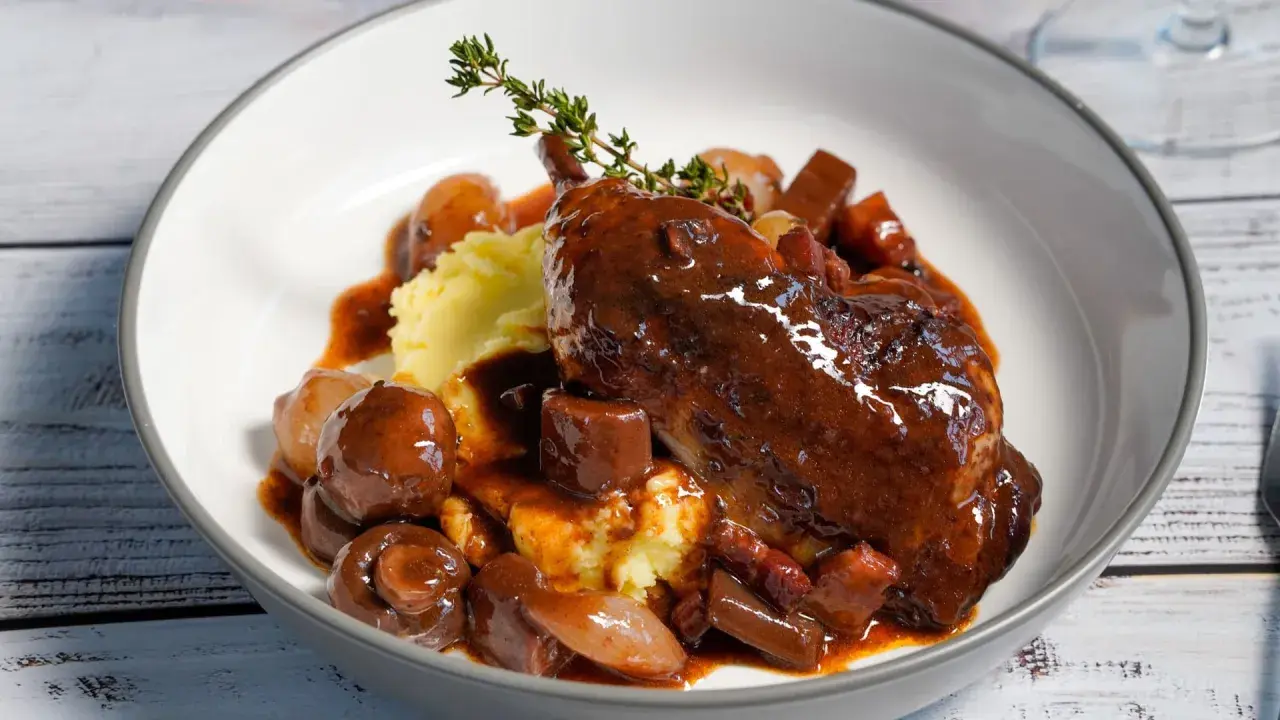 Ein authentisches Coq au Vin Rezept Original: zartes Hähnchen in Rotwein, serviert mit Kartoffelpüree, Speck und glasierten Zwiebeln.