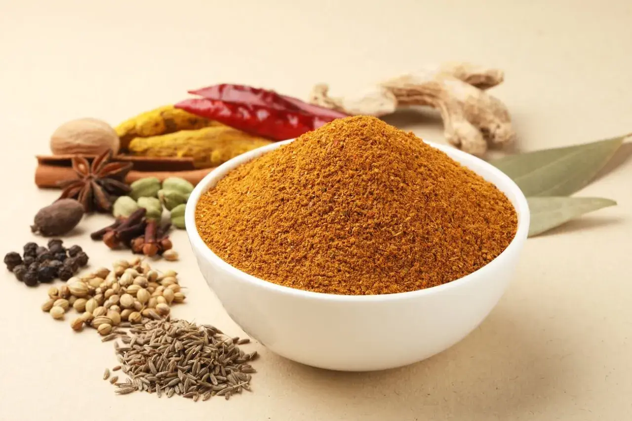 Eine Schüssel mit dem würzigen Pulver, das ist garam masala, umgeben von ganzen Gewürzen wie Zimt, Sternanis, Kardamom, Nelken, Pfeffer, Koriander, Kreuzkümmel, Ingwer und Chili.