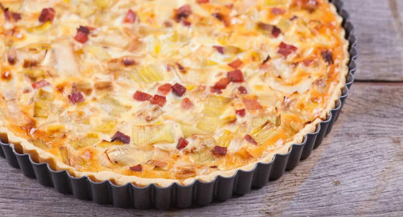 Eine herzhafte Quiche Lorraine mit Lauch und Speck in einer Metallform auf Holzhintergrund.