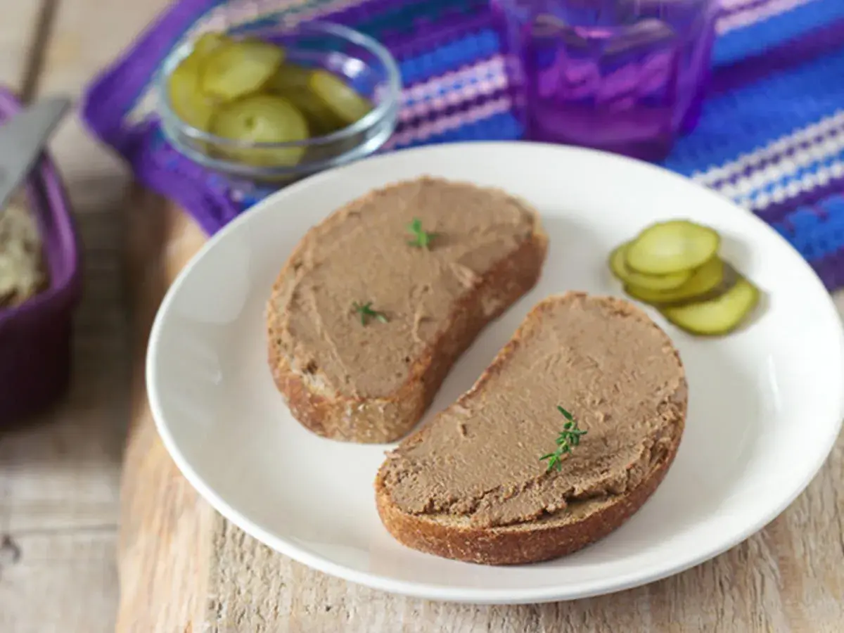 Zwei Scheiben Brot mit selbstgemachter Leberwurst, garniert mit Kräutern, dazu Gewürzgurken und ein lila Glas.