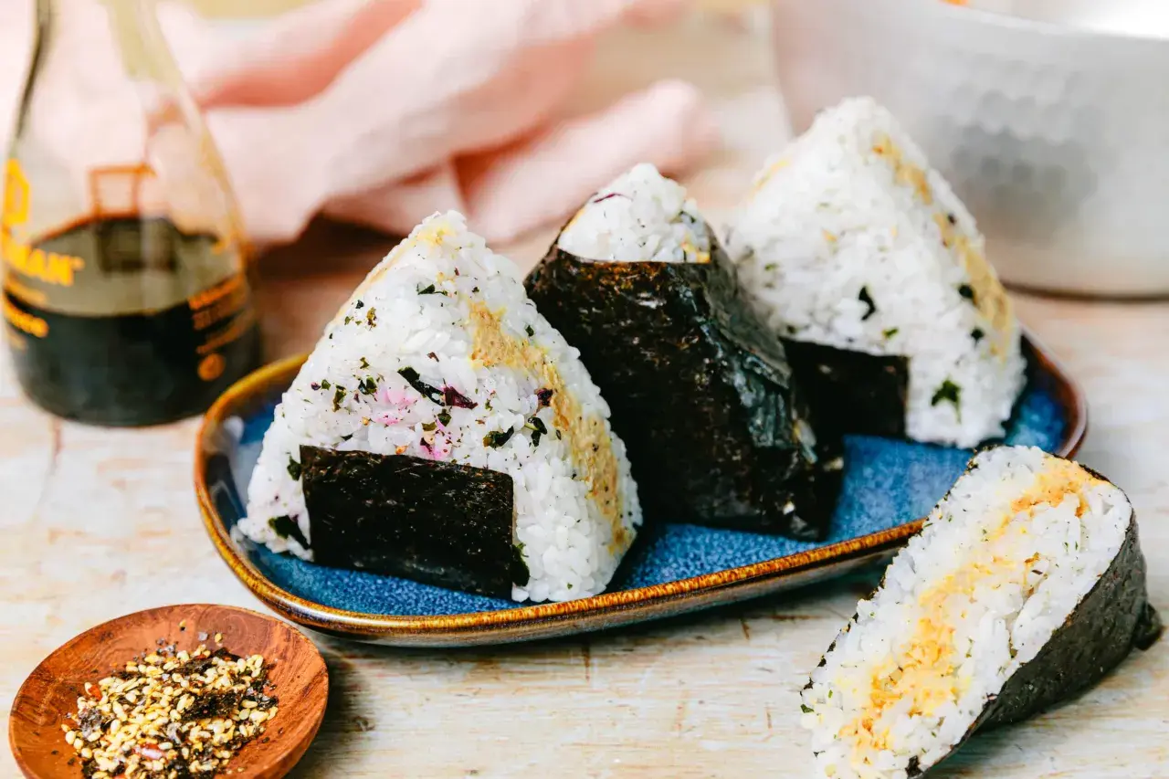 Drei dreieckige Onigiri mit Thunfisch-Mayo-Füllung, umhüllt von Nori-Algen, auf einem blauen Teller. Daneben eine Flasche Sojasauce und eine Schale mit Gewürzen.