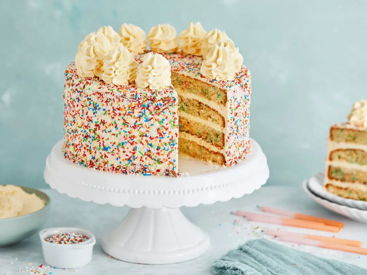 Eine leckere Torte mit weißem Zuckerguss und bunten Streuseln, gekrönt mit Sahnetuffs. Ein Stück ist angeschnitten und zeigt die saftigen Schichten.