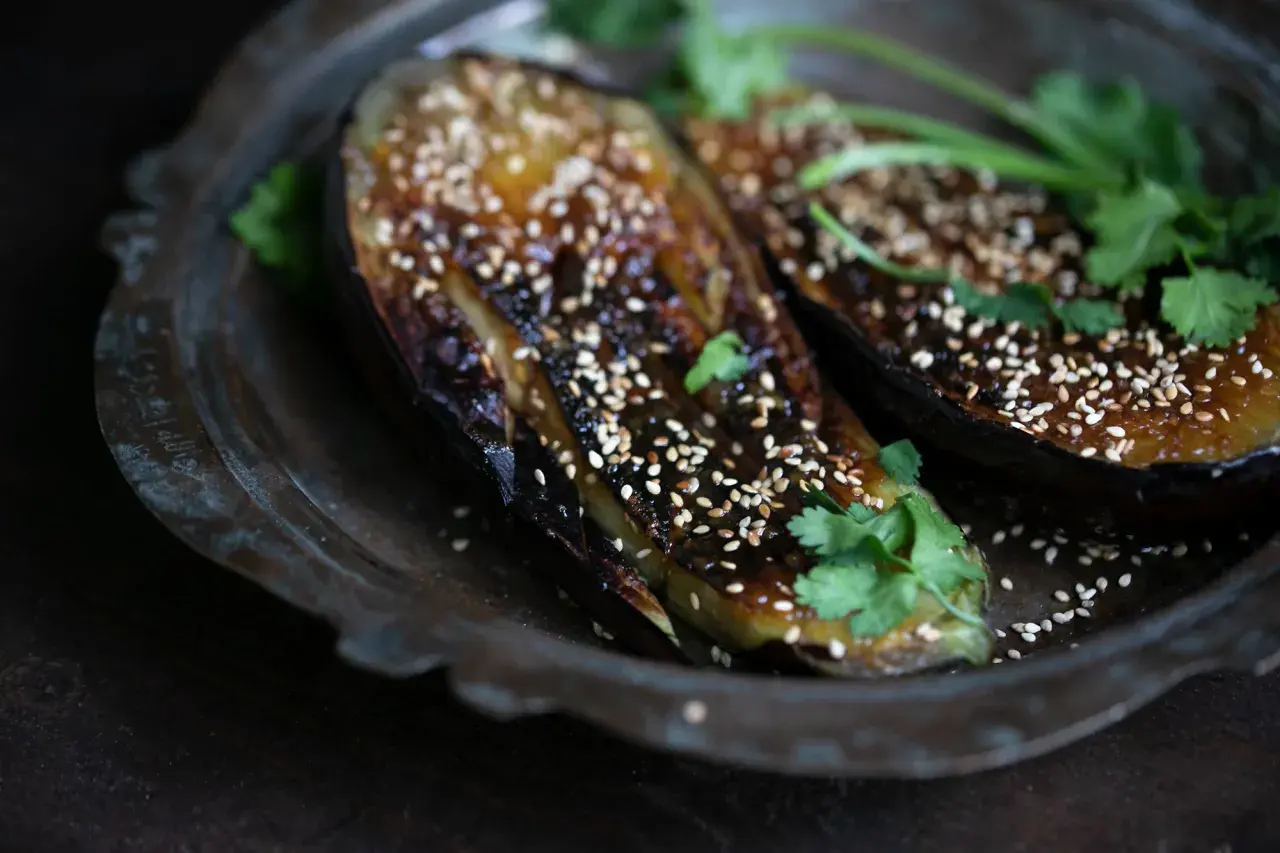 Zwei Hälften von gegrillter Miso Aubergine, bestreut mit Sesam und garniert mit Koriander, auf einem rustikalen Teller.