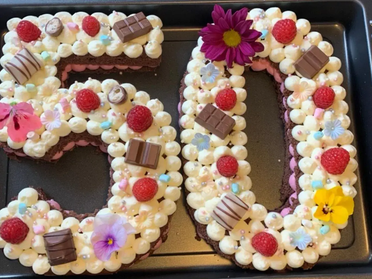 Ein aufwendig dekorierter Number Cake in Form der Zahl 50, verziert mit Sahnetuffs, Himbeeren, Schokolade und Blumen.