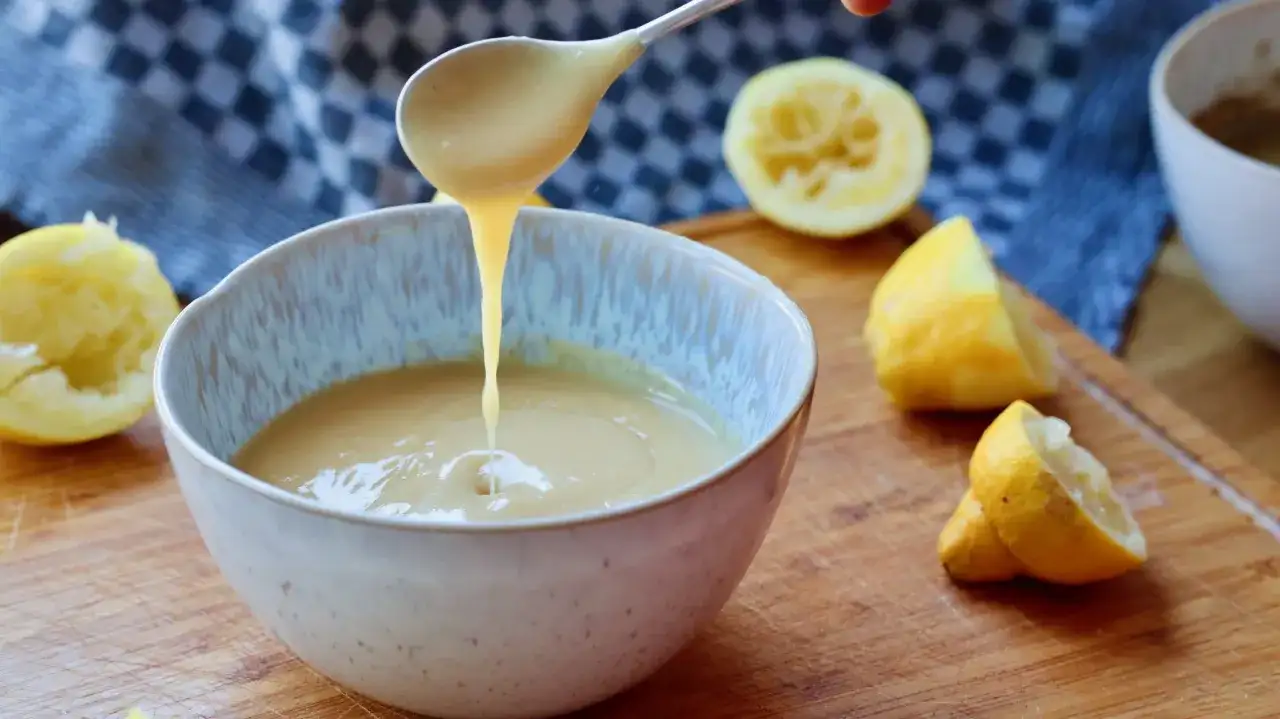 Ein Löffel cremigen Lemon Curd tropft in eine Schüssel. Frische Zitronen liegen daneben.