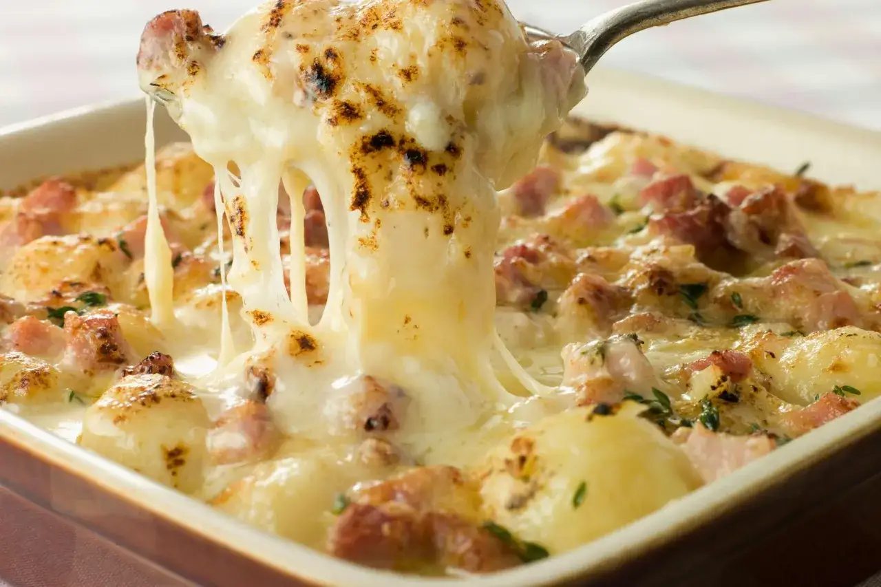 Ein L&ouml;ffel hebt eine Portion cremige Tartiflette mit geschmolzenem K&auml;se und Speck aus einer Auflaufform. Perfekt f&uuml;r ein Tartiflette Rezept!
