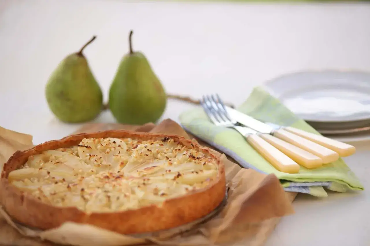 Eine k&ouml;stliche Birnen-Quiche mit N&uuml;ssen, daneben zwei frische Birnen, Besteck und Teller.