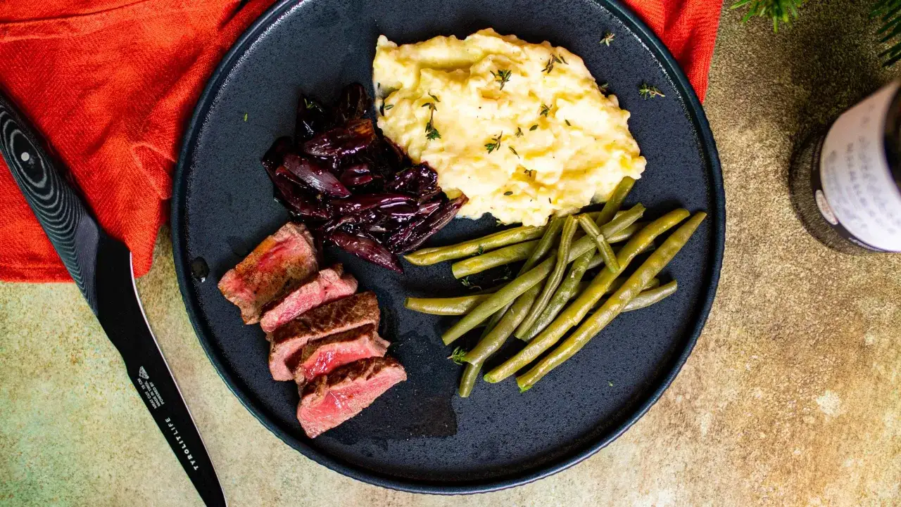 Ein perfekt gebratenes Filet de Boeuf mit Kartoffelp&uuml;ree, karamellisierten roten Zwiebeln und gr&uuml;nen Bohnen auf einem dunklen Teller.
