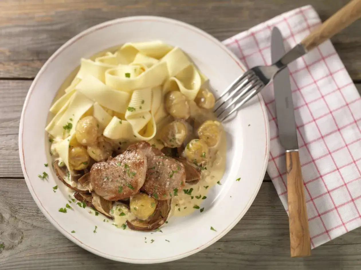 Schweinemedaillons mit Pilzen und breiten Bandnudeln in cremiger Sauce, garniert mit Petersilie. Ein Messer und eine Gabel liegen bereit.