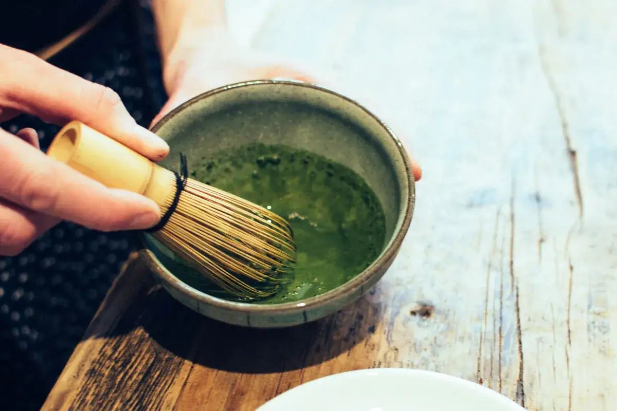 Eine Hand r&uuml;hrt mit einem Bambusbesen in einer Schale mit leuchtend gr&uuml;nem Pulver und Wasser. So wird Matcha zubereitet.