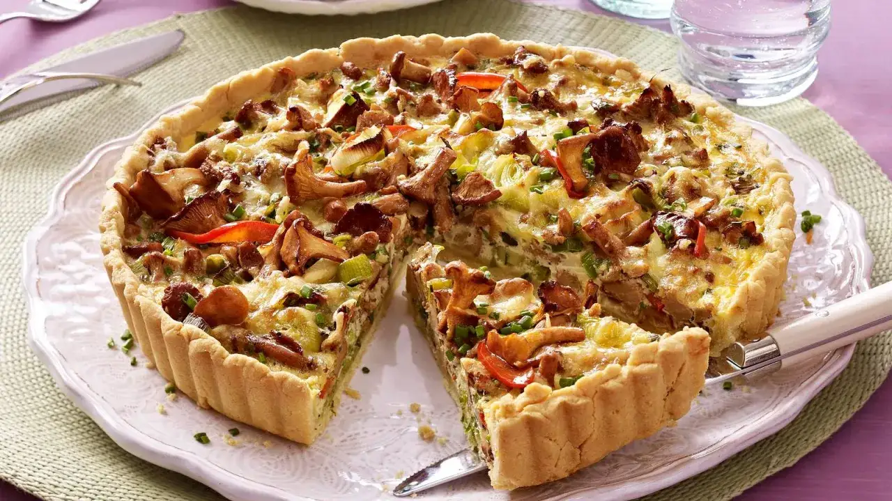 Herzhafte Quiche mit Pfifferlingen, Lauch und roten Paprikastreifen, goldbraun gebacken. Ein Stück ist bereits herausgeschnitten.