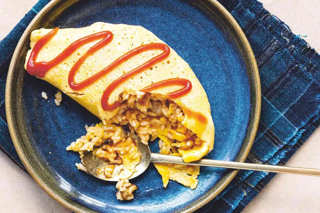 Omurice Rezept: Ein goldgelbes Omelett umhüllt herzhaften gebratenen Reis, garniert mit Ketchup. Ein Löffel zeigt die köstliche Füllung.