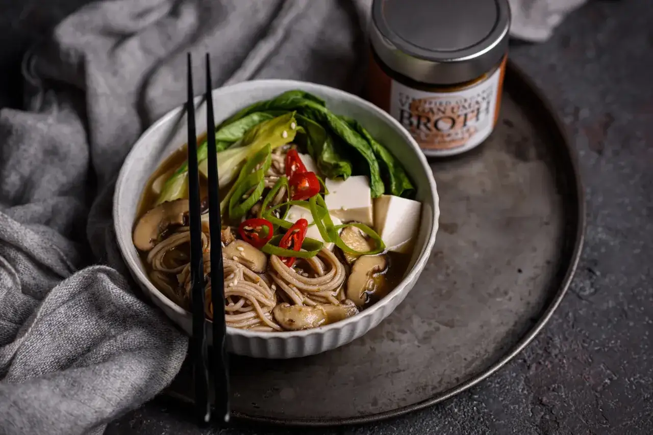 Herzhaftes, vegetarisches Ramen mit Buch-Choy, Tofu, Pilzen und Chili. Ein Glas vegane Br&uuml;he steht daneben.
