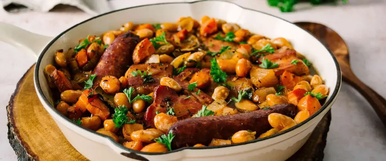 Herzhaftes Cassoulet-Rezept in einer Pfanne: weiße Bohnen, Karotten, Speck und Würstchen, garniert mit Petersilie.