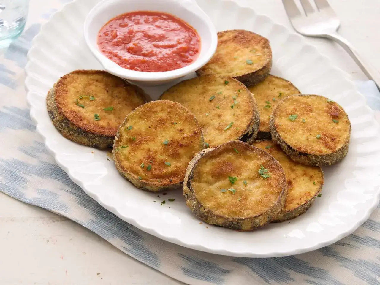 Knusprig frittierte Aubergine-Scheiben, garniert mit Petersilie, serviert mit einer Schale Tomatensauce.