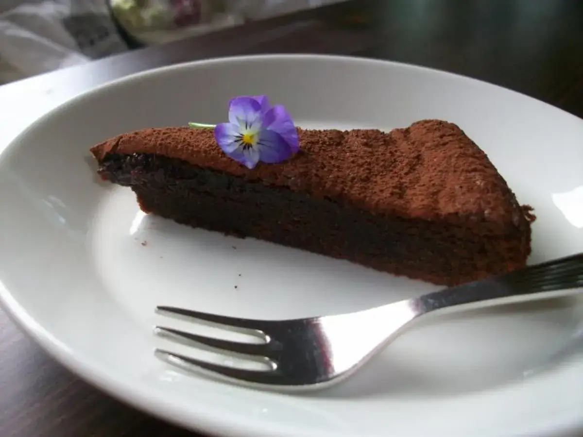Ein St&uuml;ck schokokuchen mit weichem kern, best&auml;ubt mit Kakao und garniert mit einer kleinen lila Blume, liegt auf einem wei&szlig;en Teller neben einer Gabel.