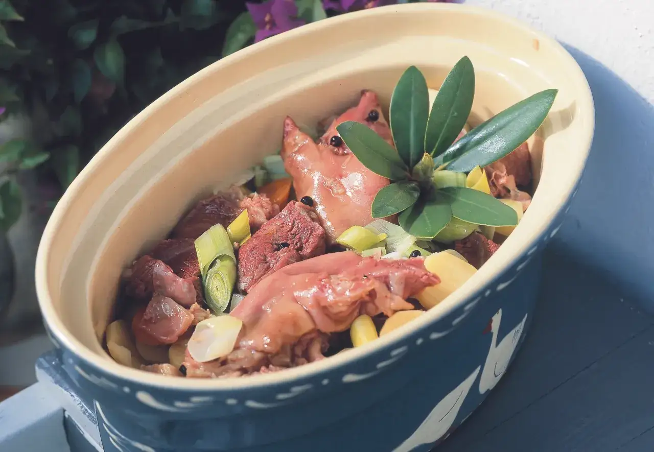 Ein traditionelles Baeckeoffe Rezept original: Fleisch, Kartoffeln und Lauch in einem Keramiktopf, garniert mit grünen Blättern.