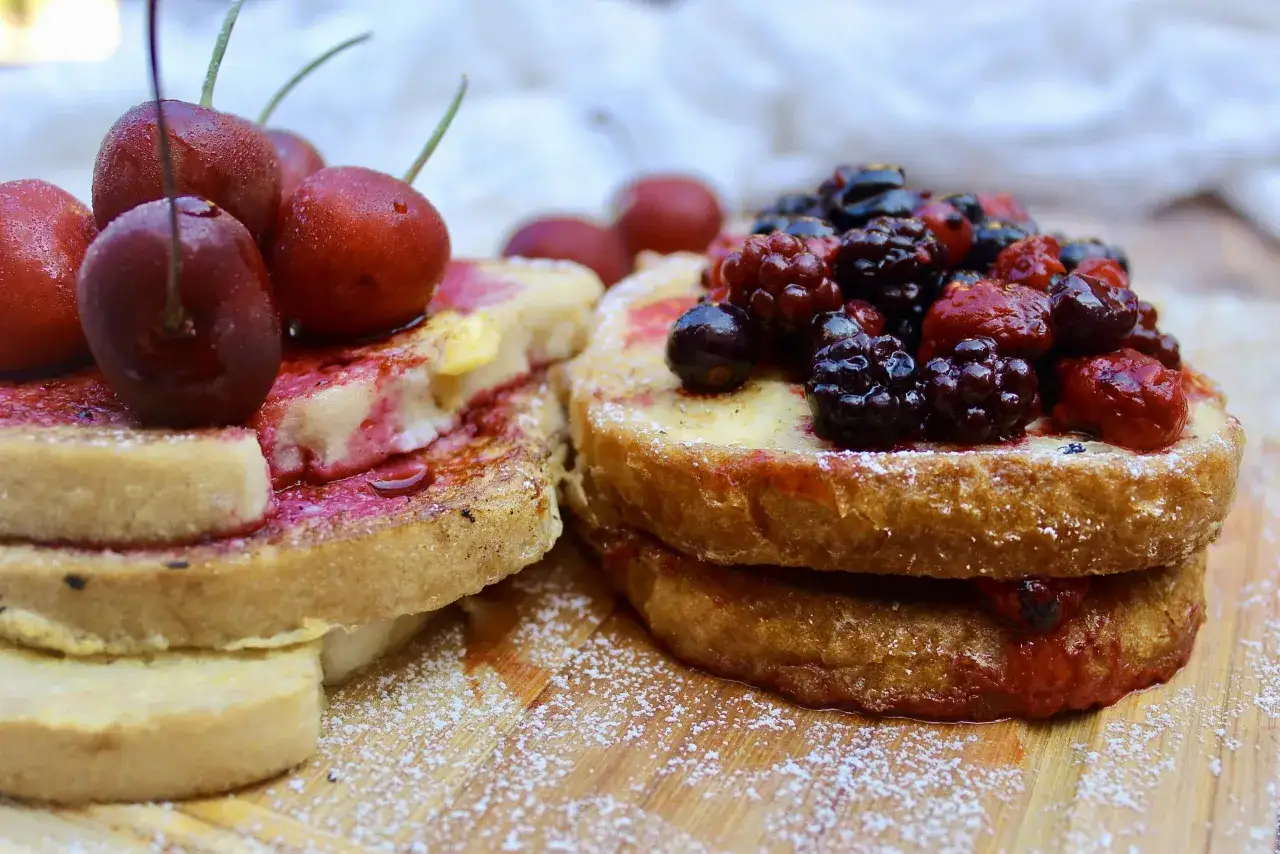 Zwei goldbraune Scheiben Pain Perdu, üppig belegt mit frischen Kirschen und Beeren, bestäubt mit Puderzucker.