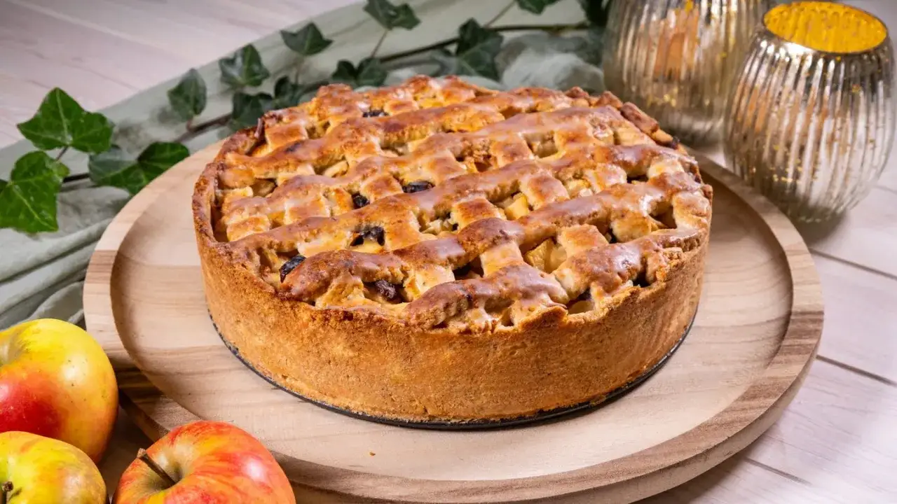 Ein goldbrauner Apfelkuchen mit Gittermuster, perfekt für ein Apfelkuchen Rezept. Daneben liegen frische Äpfel.