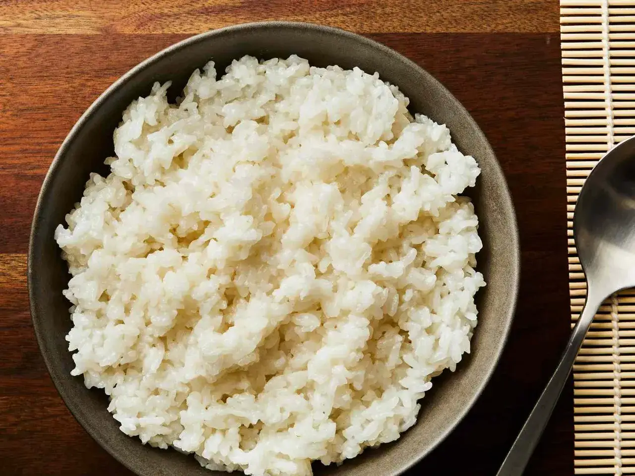 Eine Sch&uuml;ssel mit dampfendem, perfekt gekochtem Sushi Reis, bereit f&uuml;r die Zubereitung von Sushi. Daneben ein L&ouml;ffel und eine Bambusmatte.