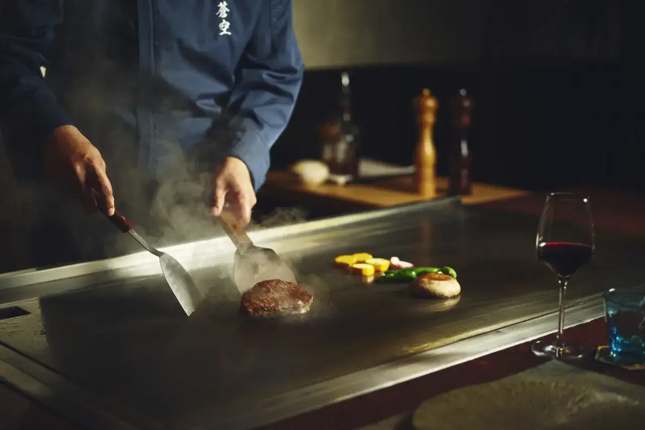 Ein Koch bereitet ein Steak und Gem&uuml;se auf einer hei&szlig;en Platte zu. Das ist Teppanyaki, eine japanische Kochkunst.
