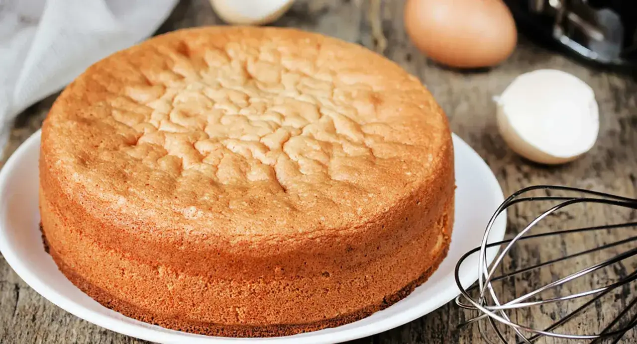 Ein goldbrauner Biskuitkuchen auf einem Teller, daneben Eier und ein Schneebesen. Perfekte Biskuit Backzeit und Temperatur sind entscheidend für dieses Ergebnis.