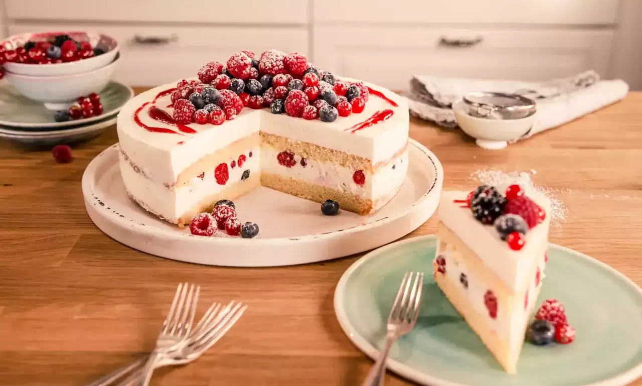 Eine leckere Torte mit Sahne und frischen Beeren, wie Himbeeren und Blaubeeren, verziert. Ein Stück ist bereits angeschnitten und auf einem Teller angerichtet.