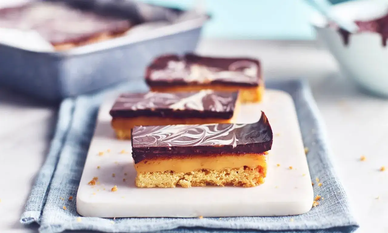 Drei saftige Millionaire Shortbread-Riegel mit Schokoladenglasur und Karamellfüllung auf einem Marmorbrett.