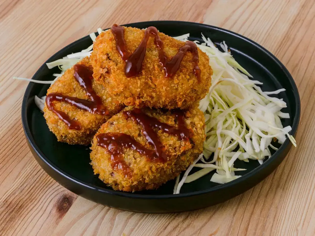 Drei knusprige japanische Kroketten mit Sauce und fein geschnittenem Kohl auf einem Teller.