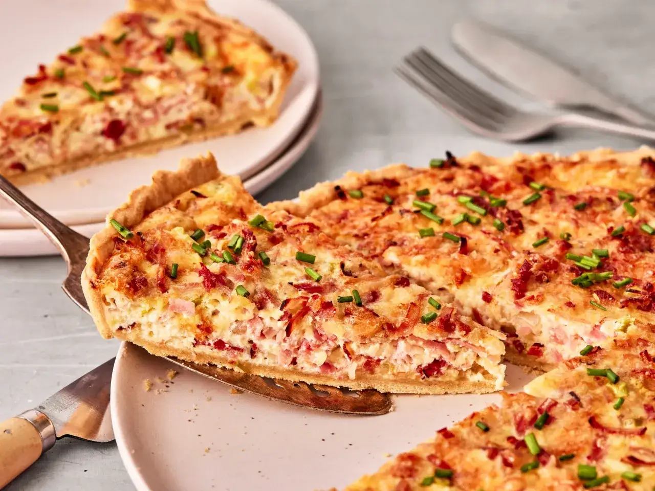 Ein Stück Quiche Lorraine wird aus einer Tarte gehoben. Darauf sind Speck und Schnittlauch zu sehen.