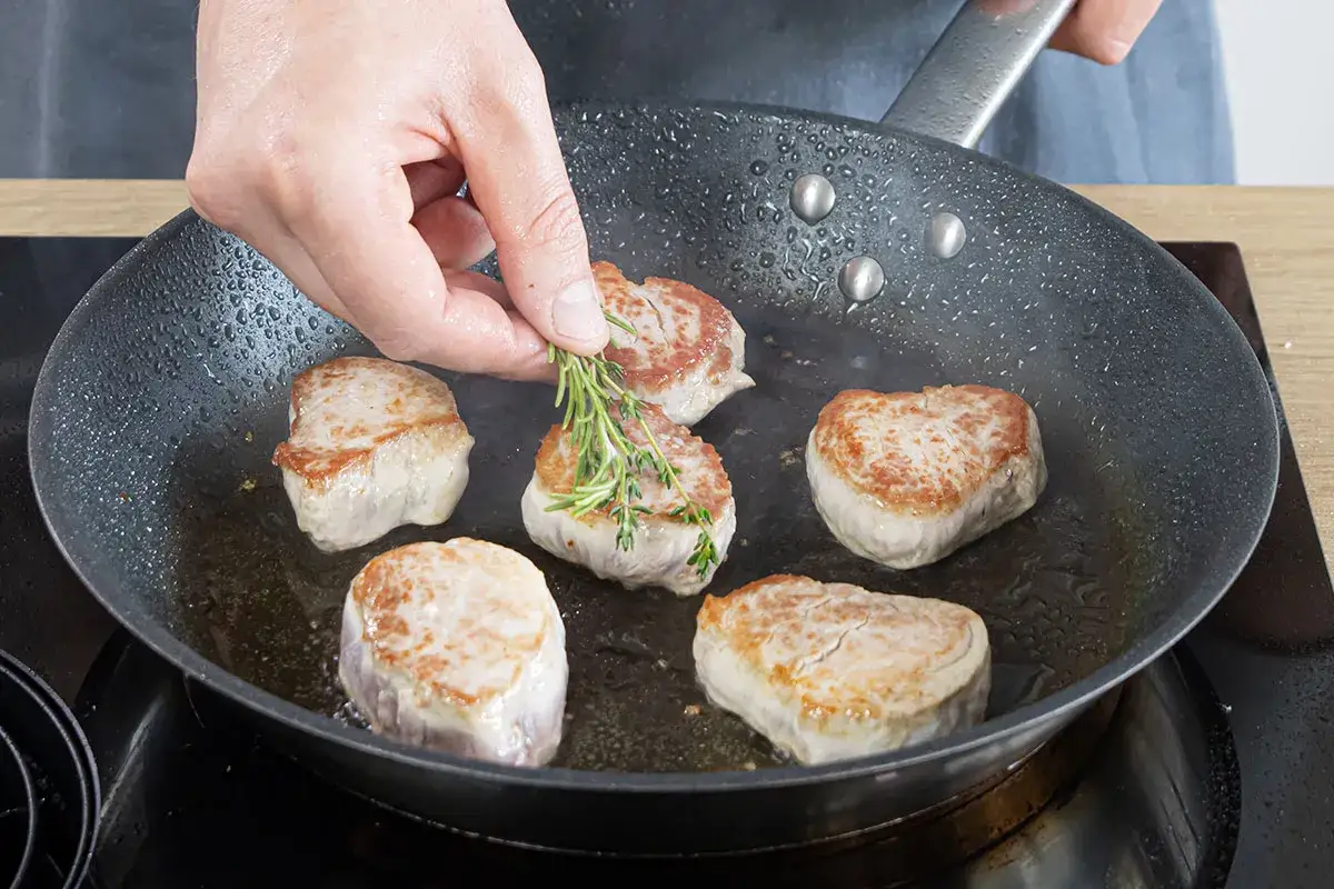 Zarte Schweinemedaillons braten in der Pfanne. Ein Rosmarinzweig wird hinzugefügt, um das Aroma zu verfeinern – ein köstliches Schweinemedaillons Rezept.