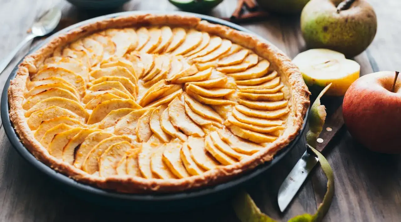 Ein goldbraunes Apfeltarte-Rezept mit d&uuml;nn geschnittenen &Auml;pfeln, arrangiert zu einem Muster. Daneben liegen frische &Auml;pfel und ein Messer.