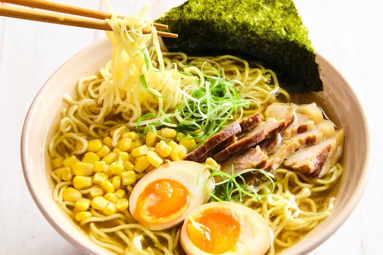 Sch&uuml;ssel mit japanischem Ramen: Nudeln, Schweinefleisch, Ei, Mais und Nori. Chopsticks heben Nudeln an.